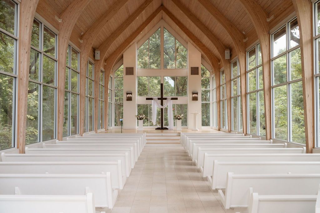 Elegant chapel interior with white pews, wood-beamed ceiling, large glass windows, and a cross at the altar.