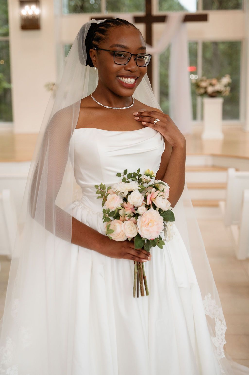 Smiling Black bride in strapless white ballgown and veil poses with blush roses in chapel.