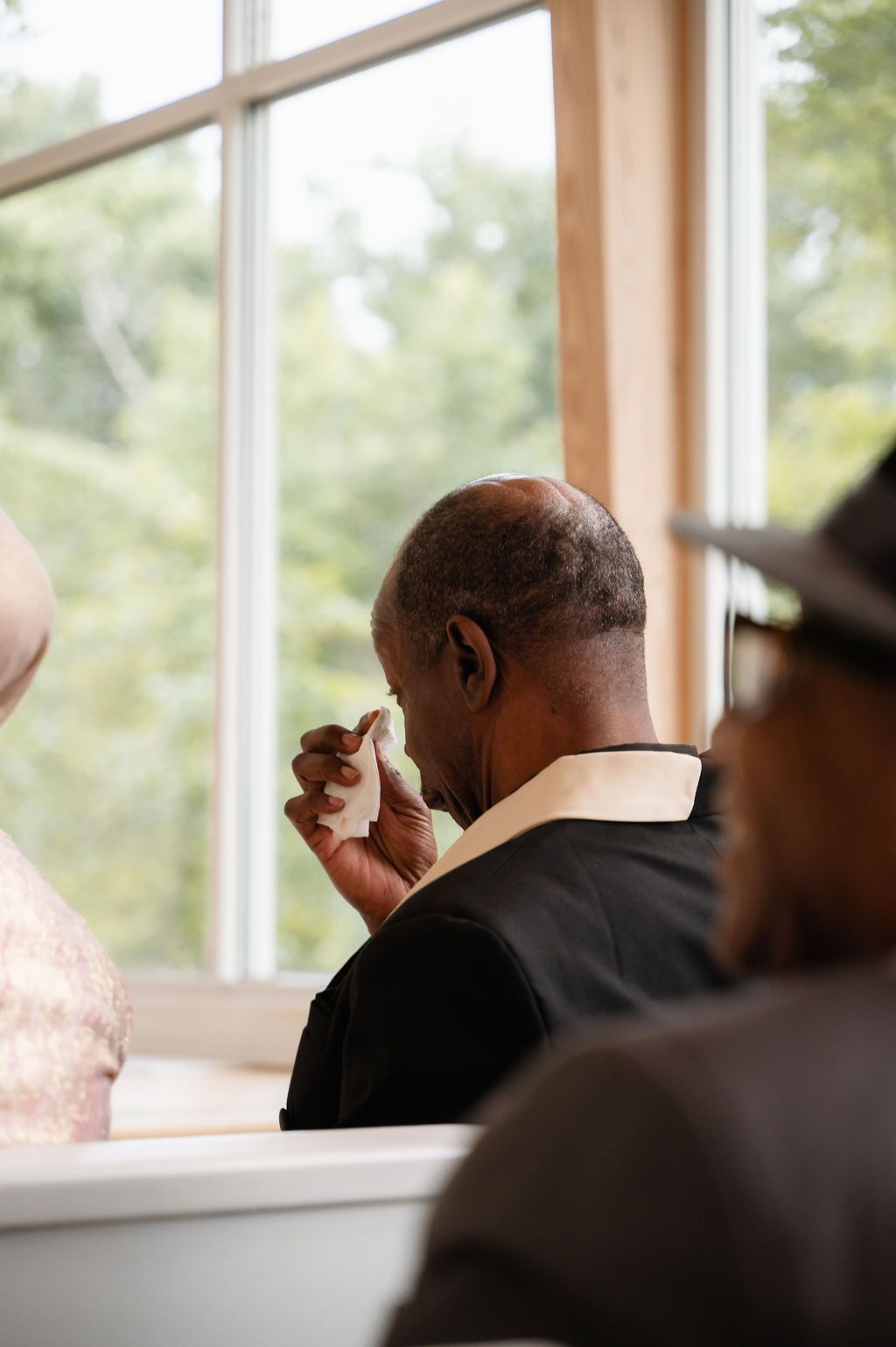 Emotional man wiping tears with handkerchief during a ceremony, seated near a bright window with greenery outside.