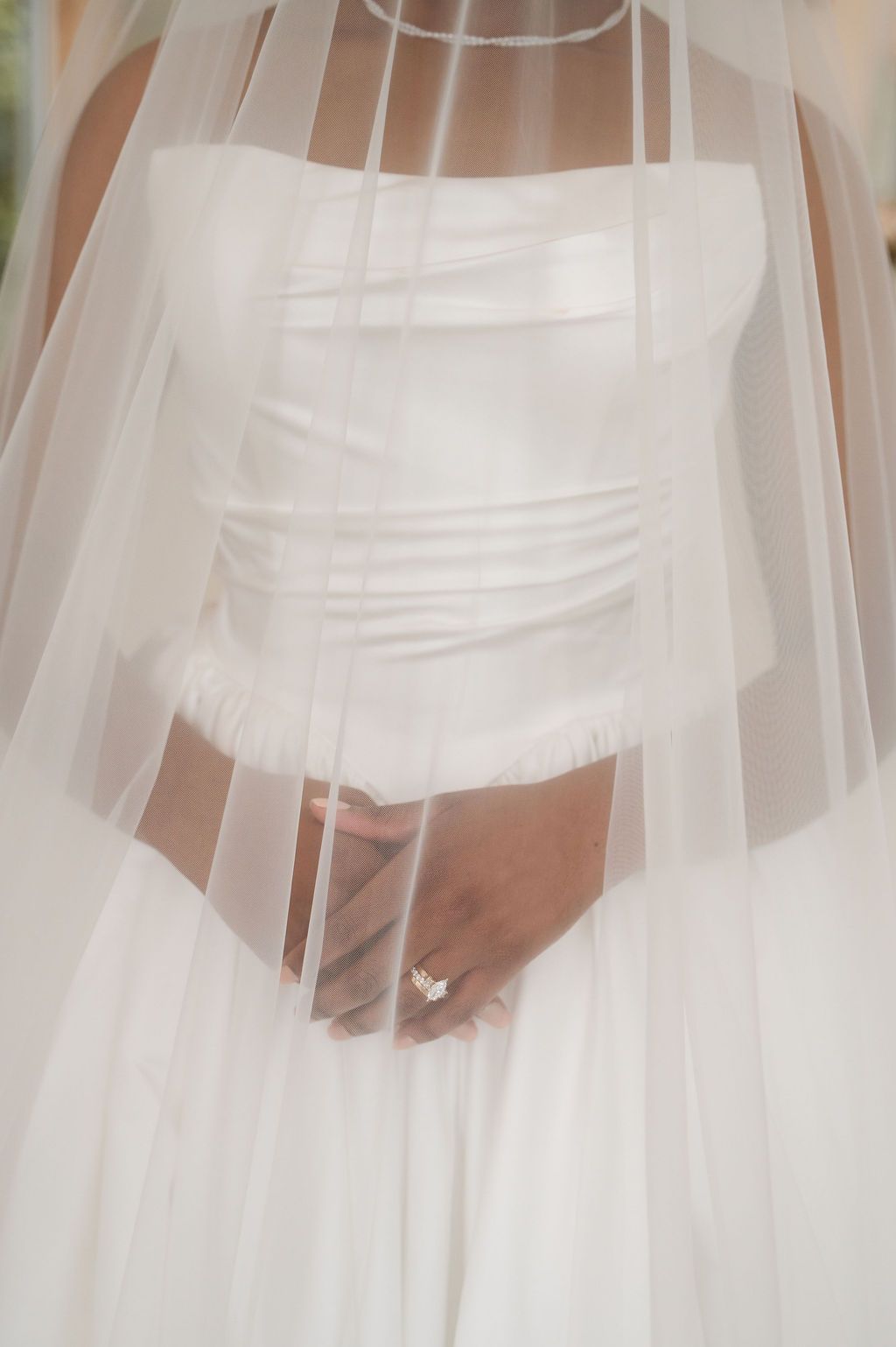Bride in strapless white wedding gown with sheer veil, hands clasped showing diamond engagement ring.