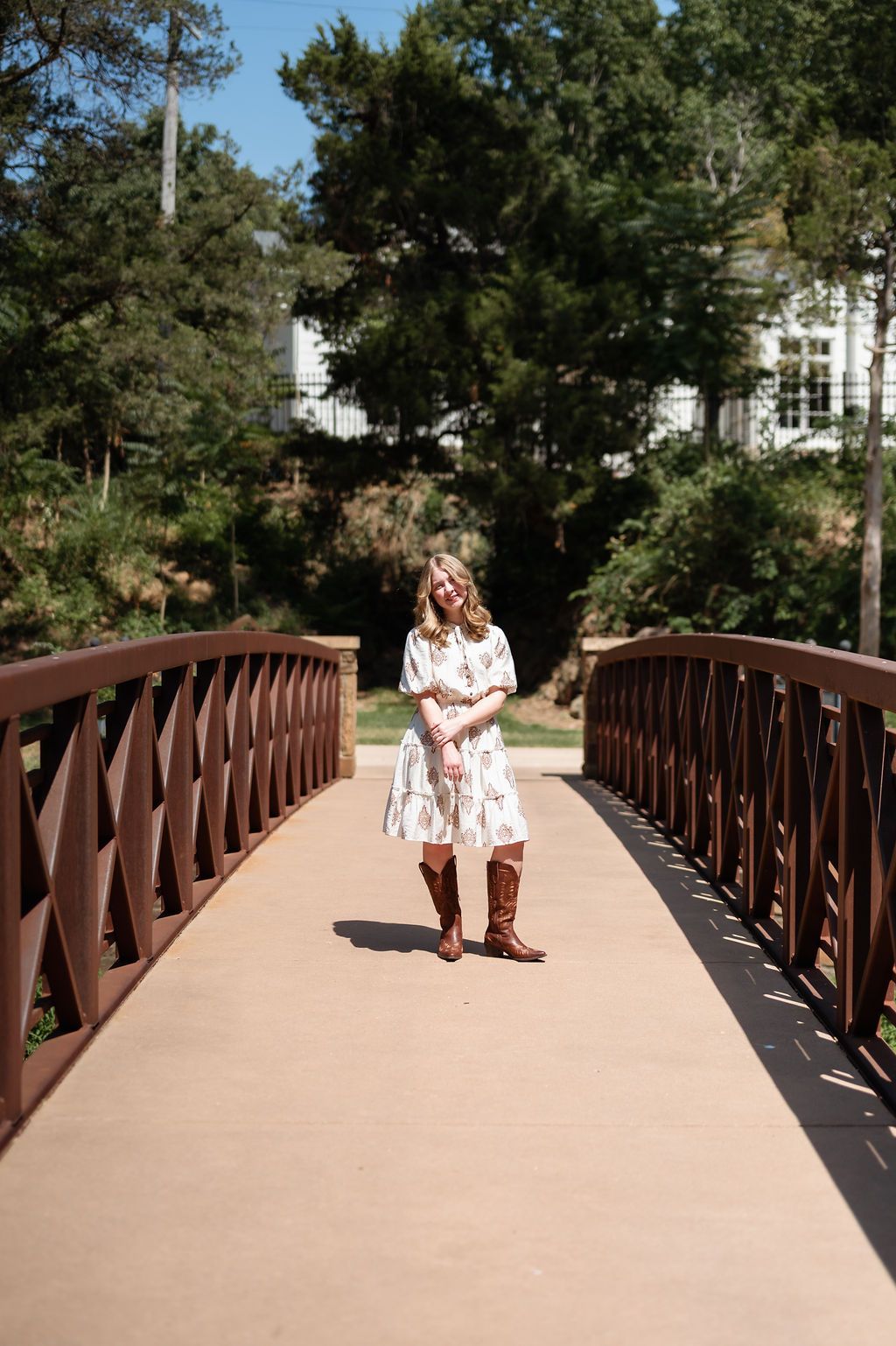 senior girl in floral dress and brown cowboy boots standing on a wooden bridge outdoors.
