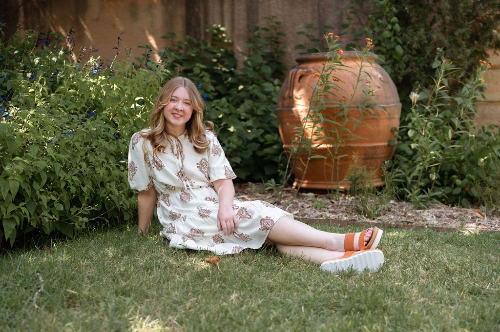 Laughing young woman in floral dress sits on grass beside large copper garden urn outdoors.