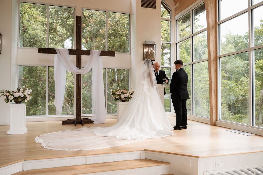 Bride and groom exchange vows at elegant chapel altar with wooden cross, white draping, and large windows overlooking lush greenery.