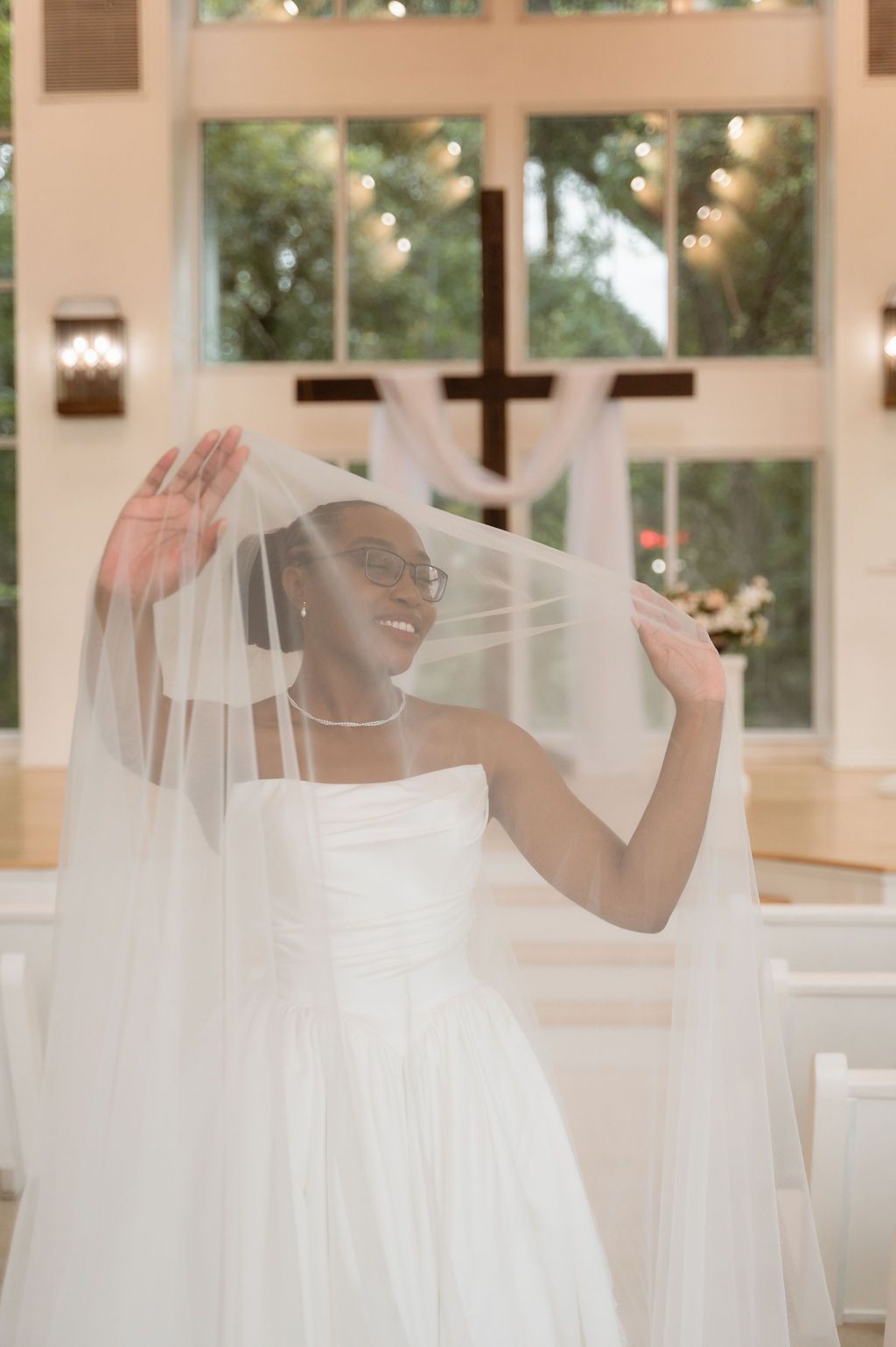 Smiling bride lifts flowing veil in bright chapel with wooden cross and large windows behind her.