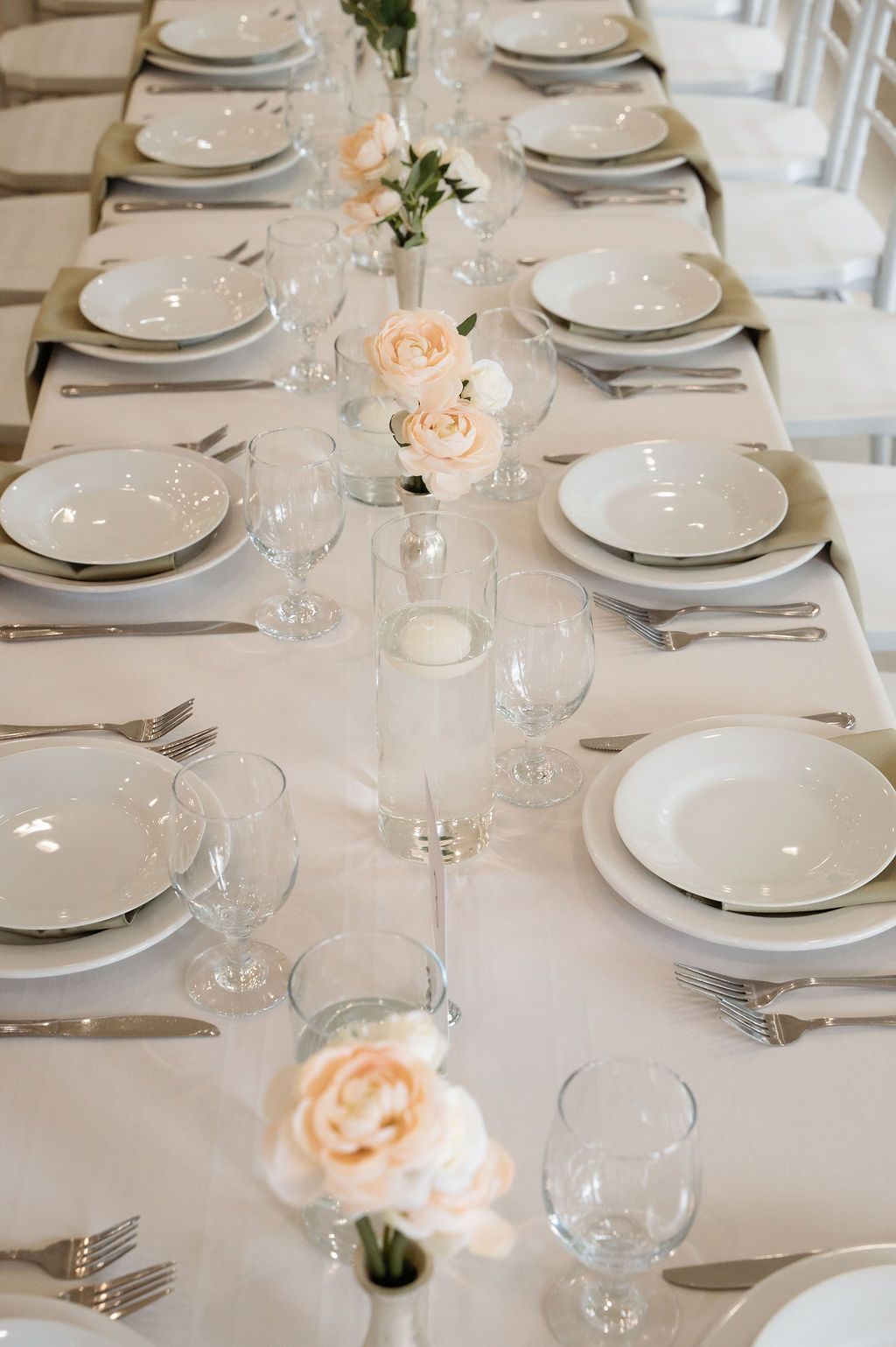 Close-up of a wedding reception table set with white plates, crystal glassware, silver cutlery, and delicate blush pink rose centerpieces.