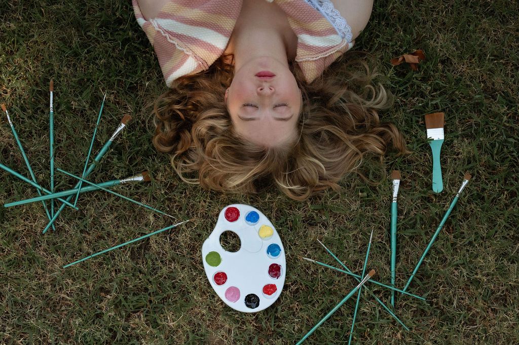 Overhead view of senior girl lying on grass surrounded by paintbrushes and a colorful paint palette at Tulsa Park
