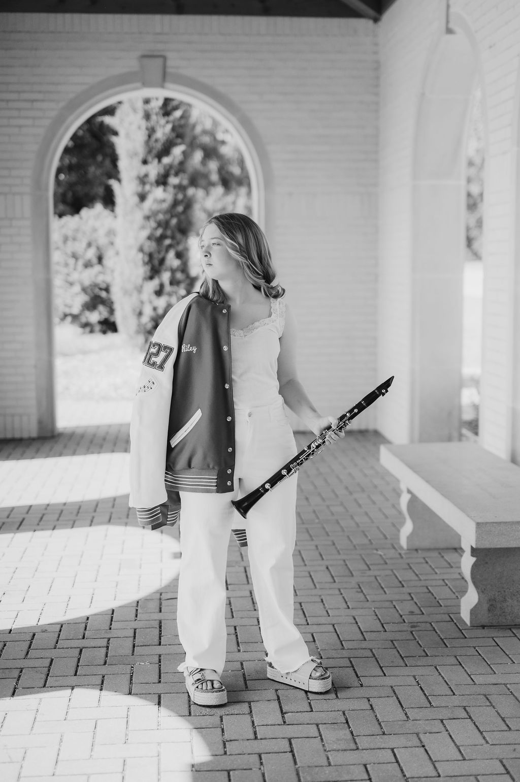 Black and white photo of a young woman holding a clarinet, wearing a varsity jacket and white pants outdoors.
