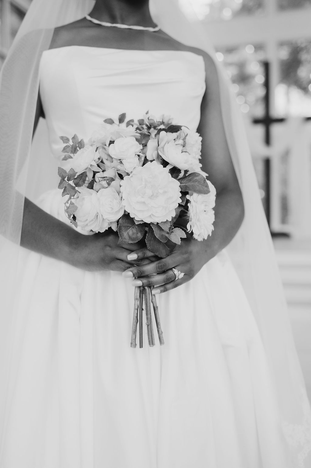 Black and white photo of bride in strapless gown holding elegant bouquet of roses and greenery.