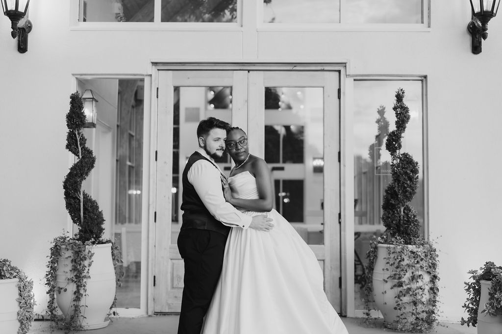 Black and white photo of an interracial bride and groom embracing in front of elegant French doors with spiral topiaries.