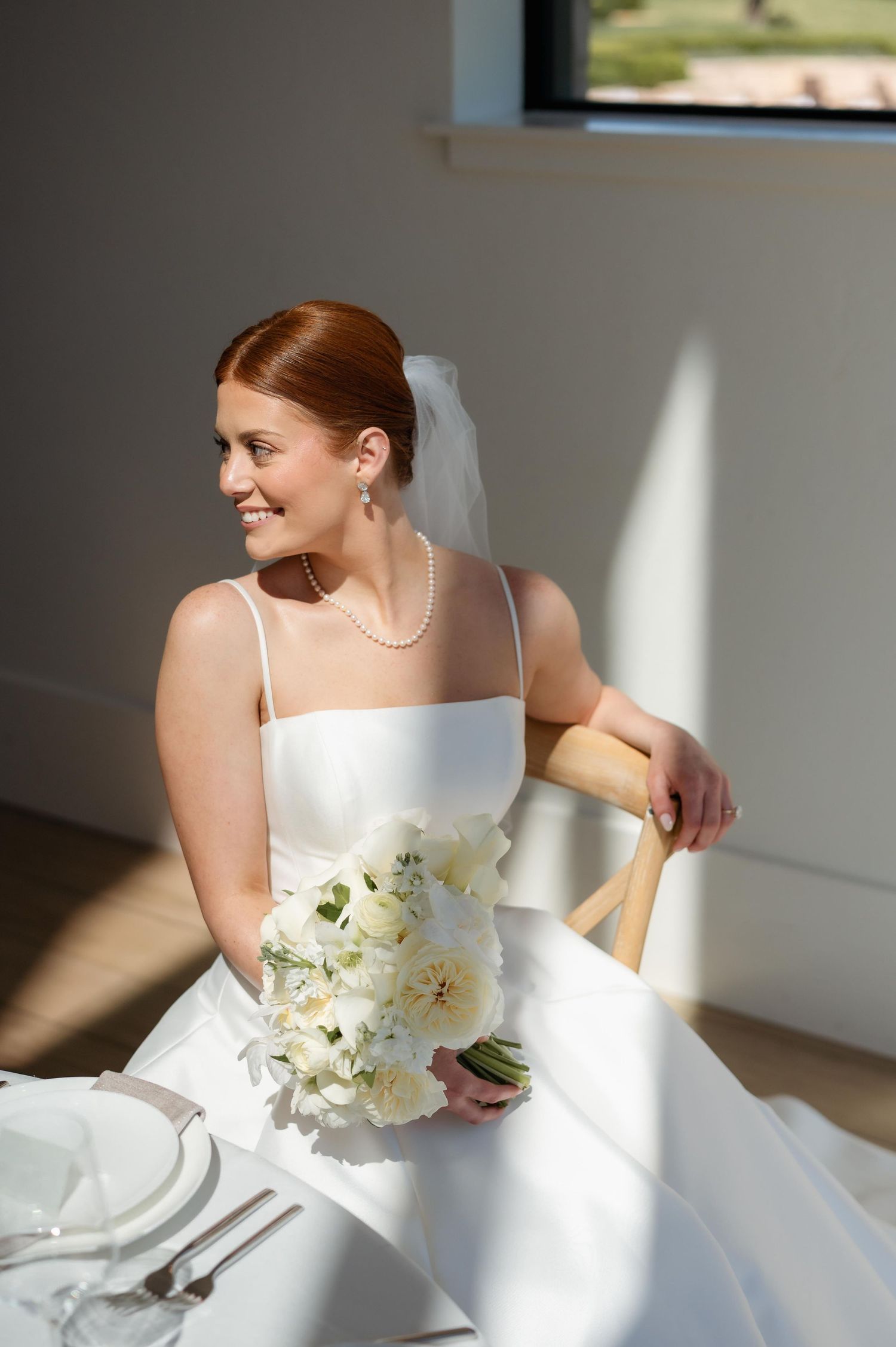 Smiling bride in white gown holding bouquet of white flowers, seated by sunlit window wearing pearl necklace and veil.
