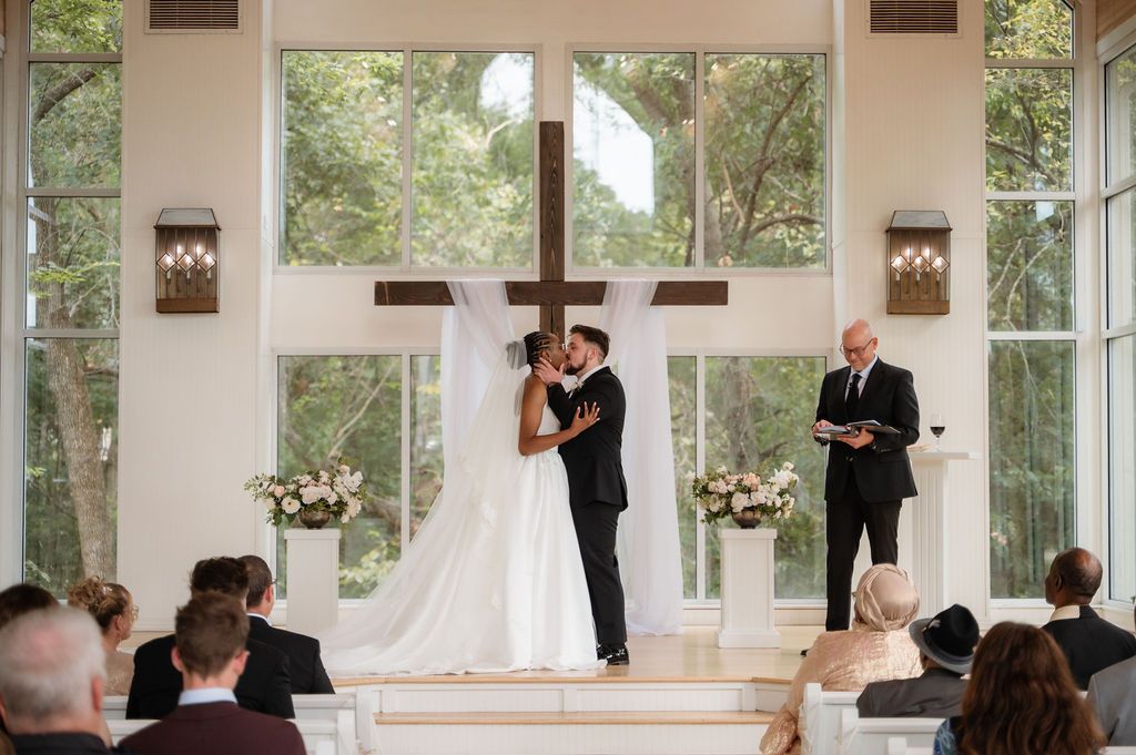 Bride and groom share first kiss at chapel altar with wooden cross and floor-to-ceiling windows during wedding ceremony.