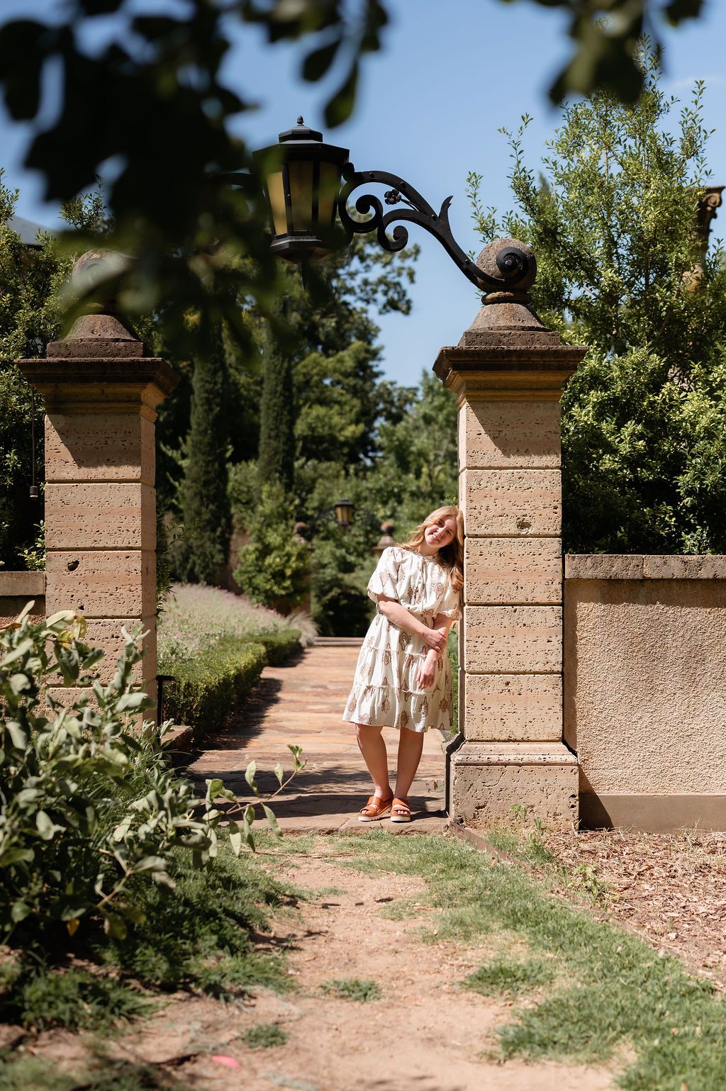 Senior Girl in floral dress leaning against stone garden gate pillar surrounded by lush greenery on a sunny day.
