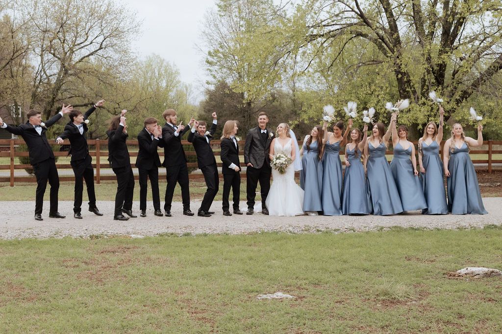 Wedding party poses outdoors, groomsmen in tuxedos and bridesmaids in blue gowns celebrating with bride and groom at Rocky Meadow Farm