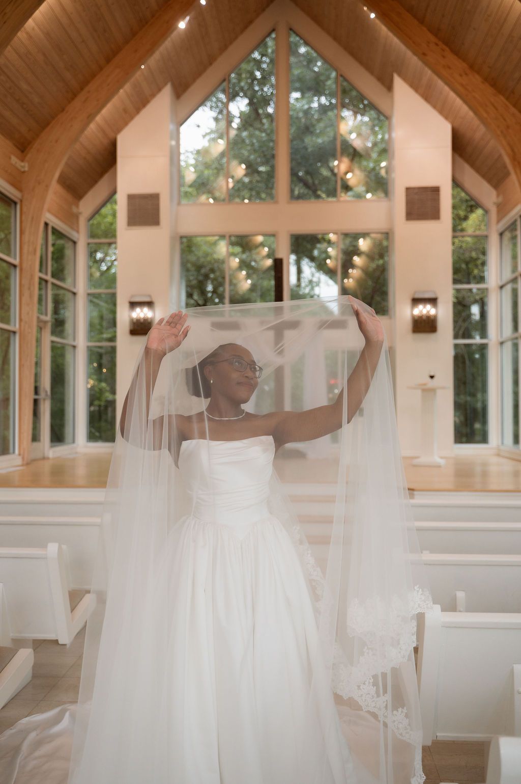 Bride in white gown lifts veil in sunlit chapel with wooden ceiling and large windows.