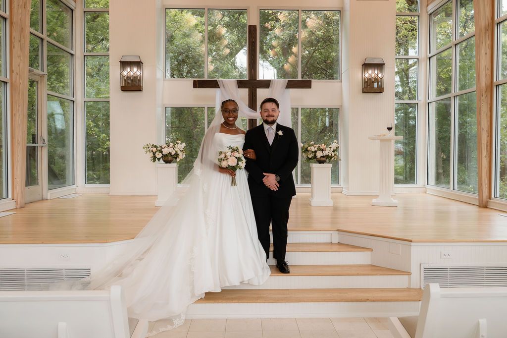 Interracial couple posing at wedding altar in elegant chapel with cross and floor-to-ceiling windows.