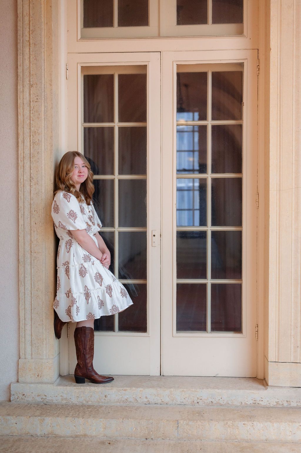 Young woman in floral dress and cowboy boots stands beside elegant French doors on a terrace.