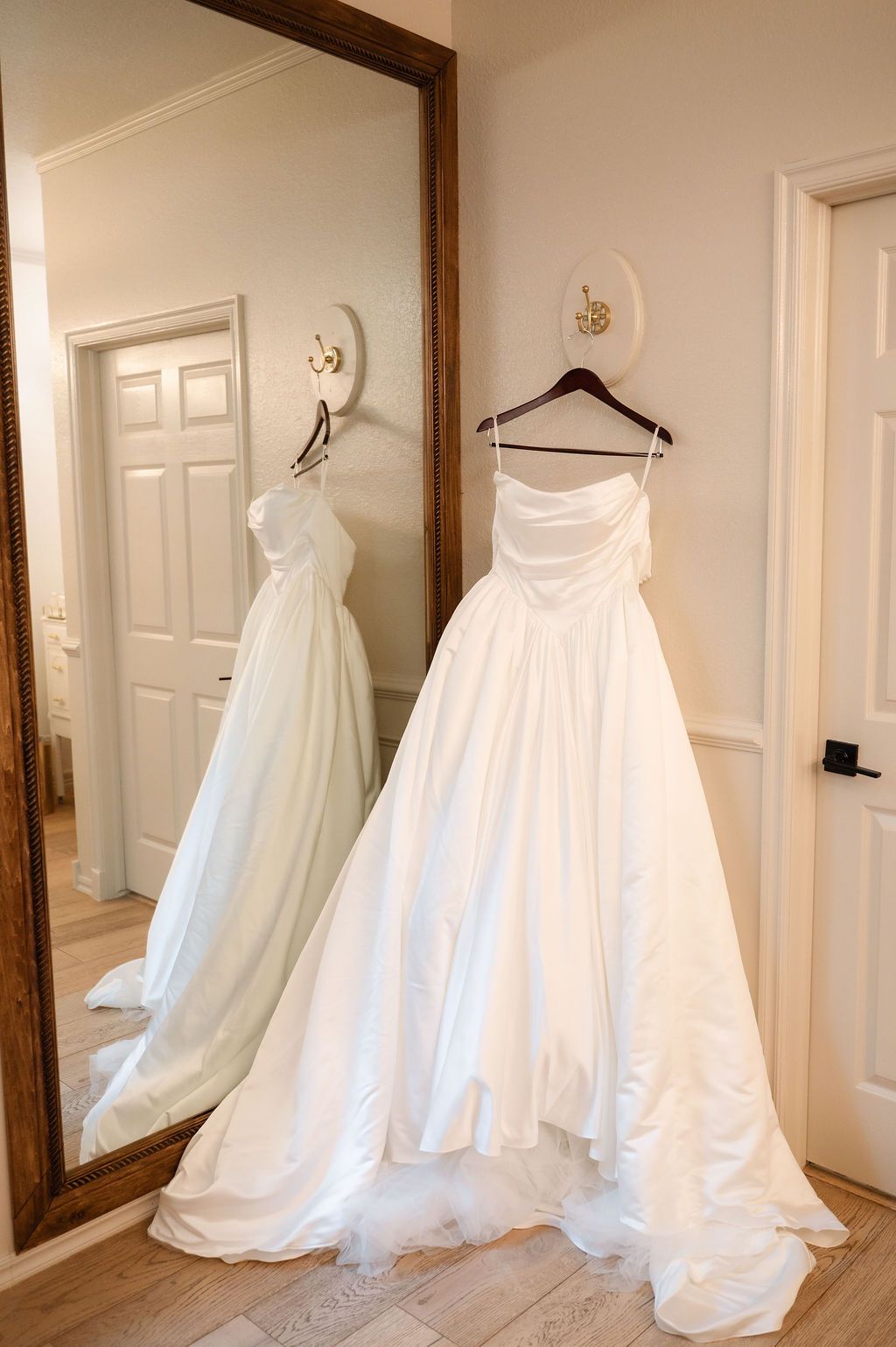 A stunning strapless white wedding ball gown with ruched bodice hanging beside a large ornate floor mirror.