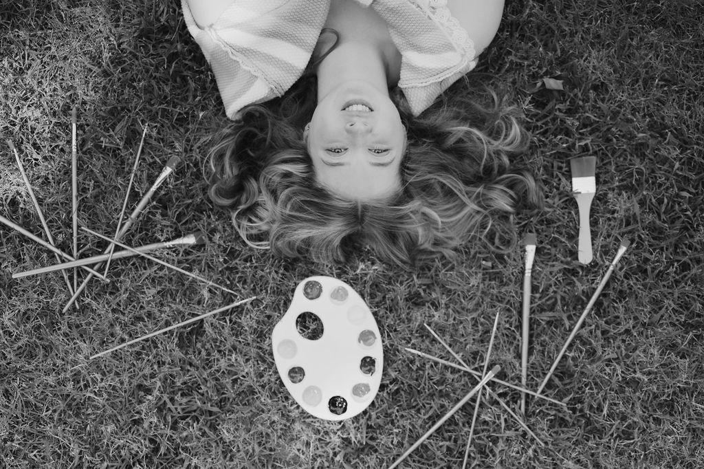 Black and white photo of a woman lying on grass surrounded by paintbrushes and an artist palette.