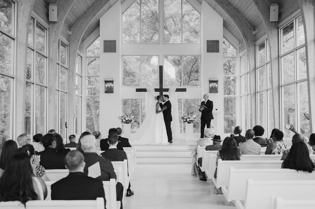 Black and white photo of a wedding ceremony in an elegant chapel with large windows and guests seated in white pews.