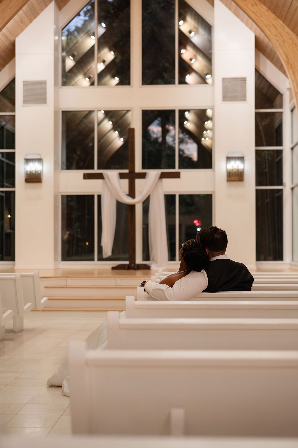 A couple shares an intimate moment seated together in a beautifully lit chapel with white pews and a wooden cross.