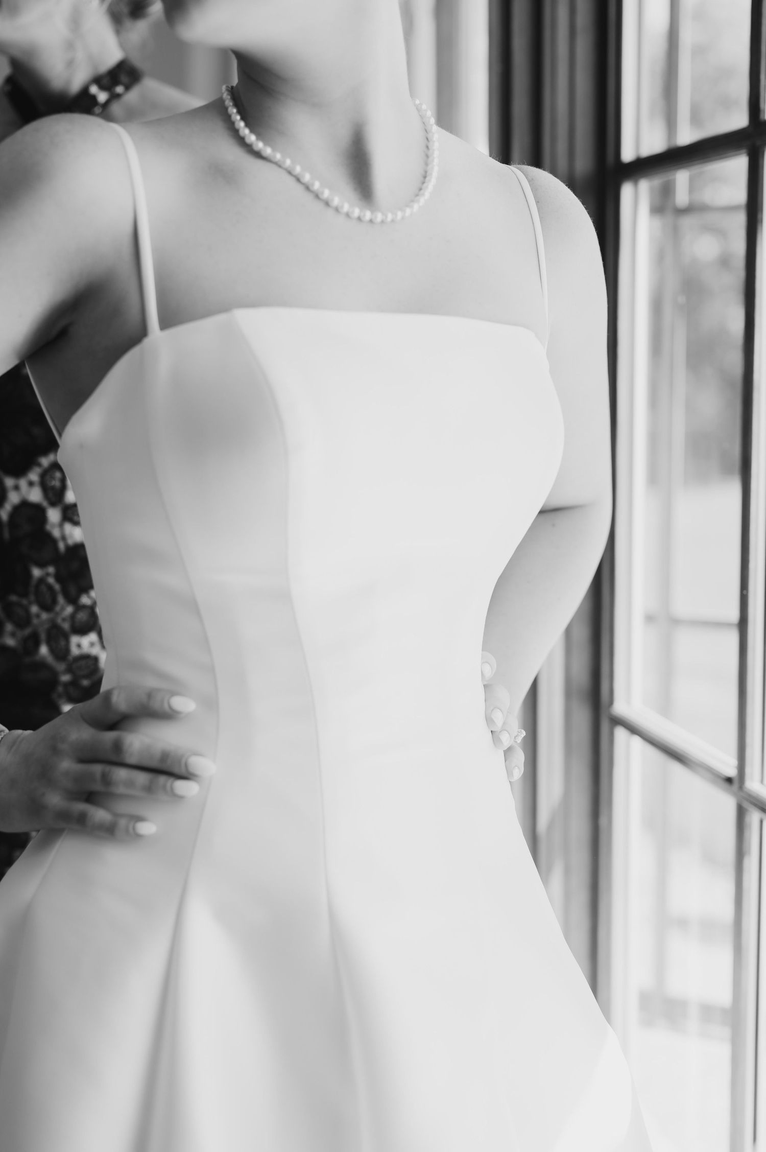 Black and white photo of a bride in a sleek white spaghetti strap wedding gown with pearl necklace near a window.