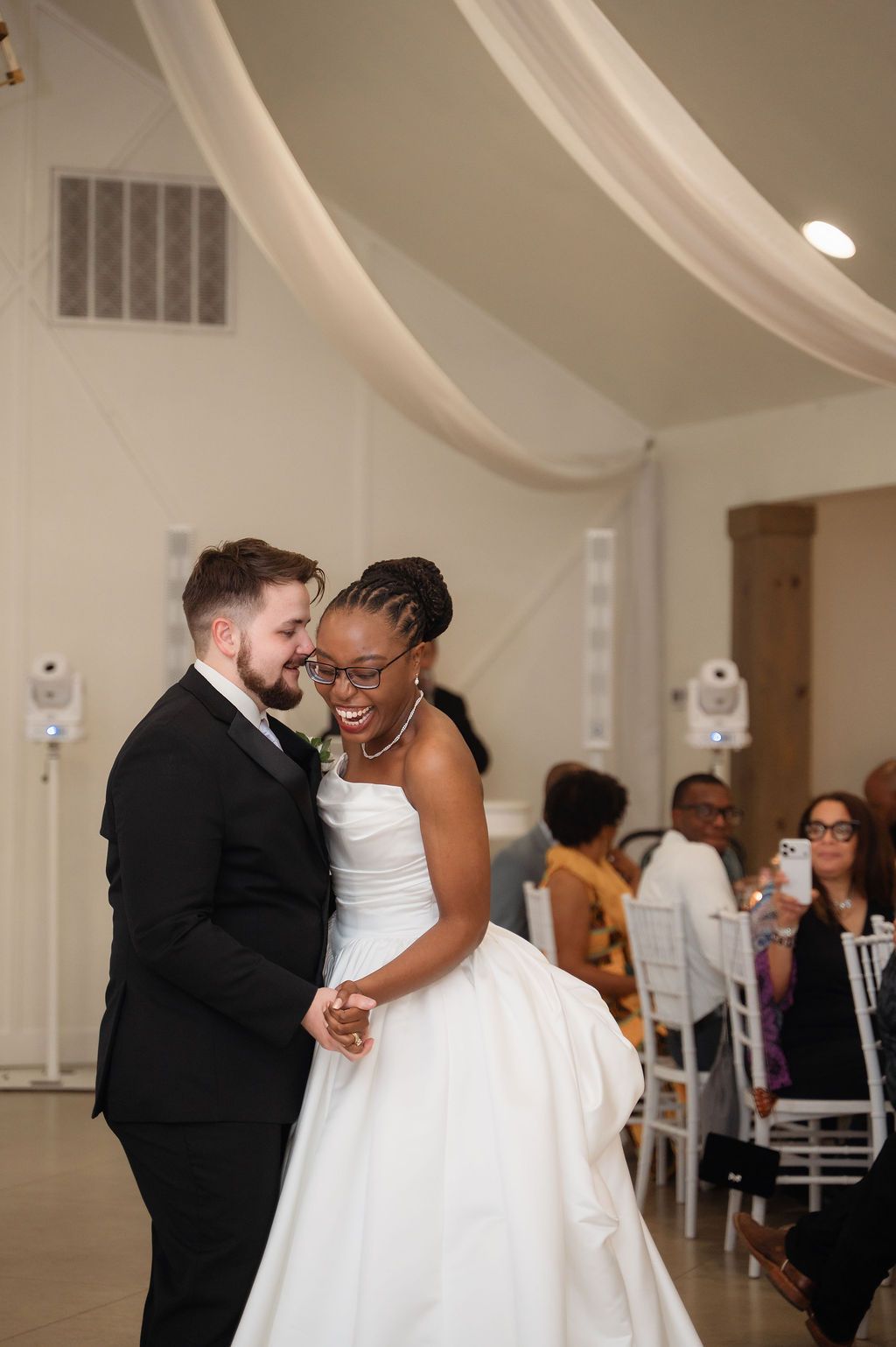Newlywed interracial couple shares joyful first dance at wedding reception, bride in white gown laughing with groom in black suit.