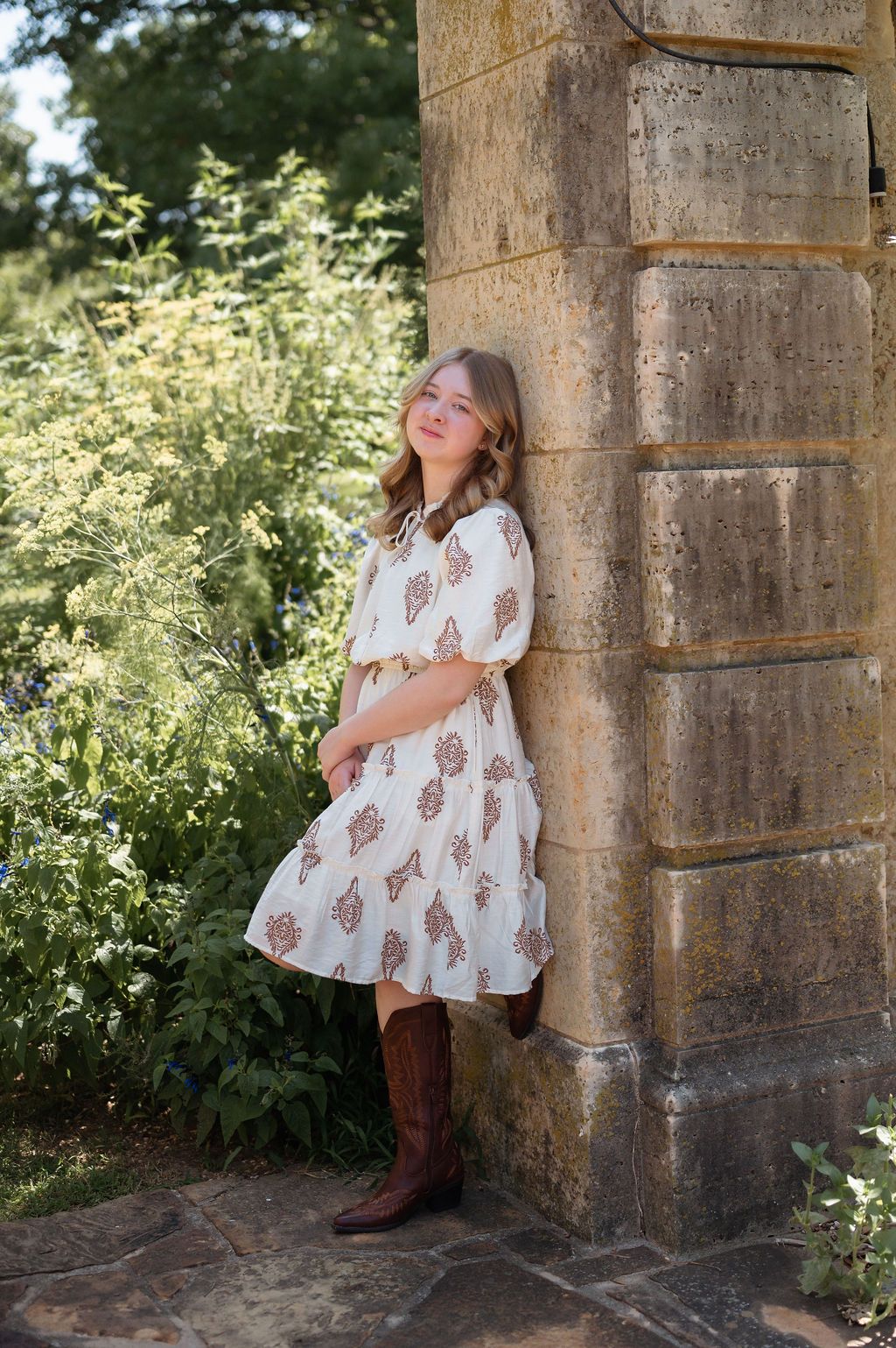 Senior girl in floral dress and brown cowboy boots leaning against stone pillar surrounded by lush green garden at Tulsa Philbrook