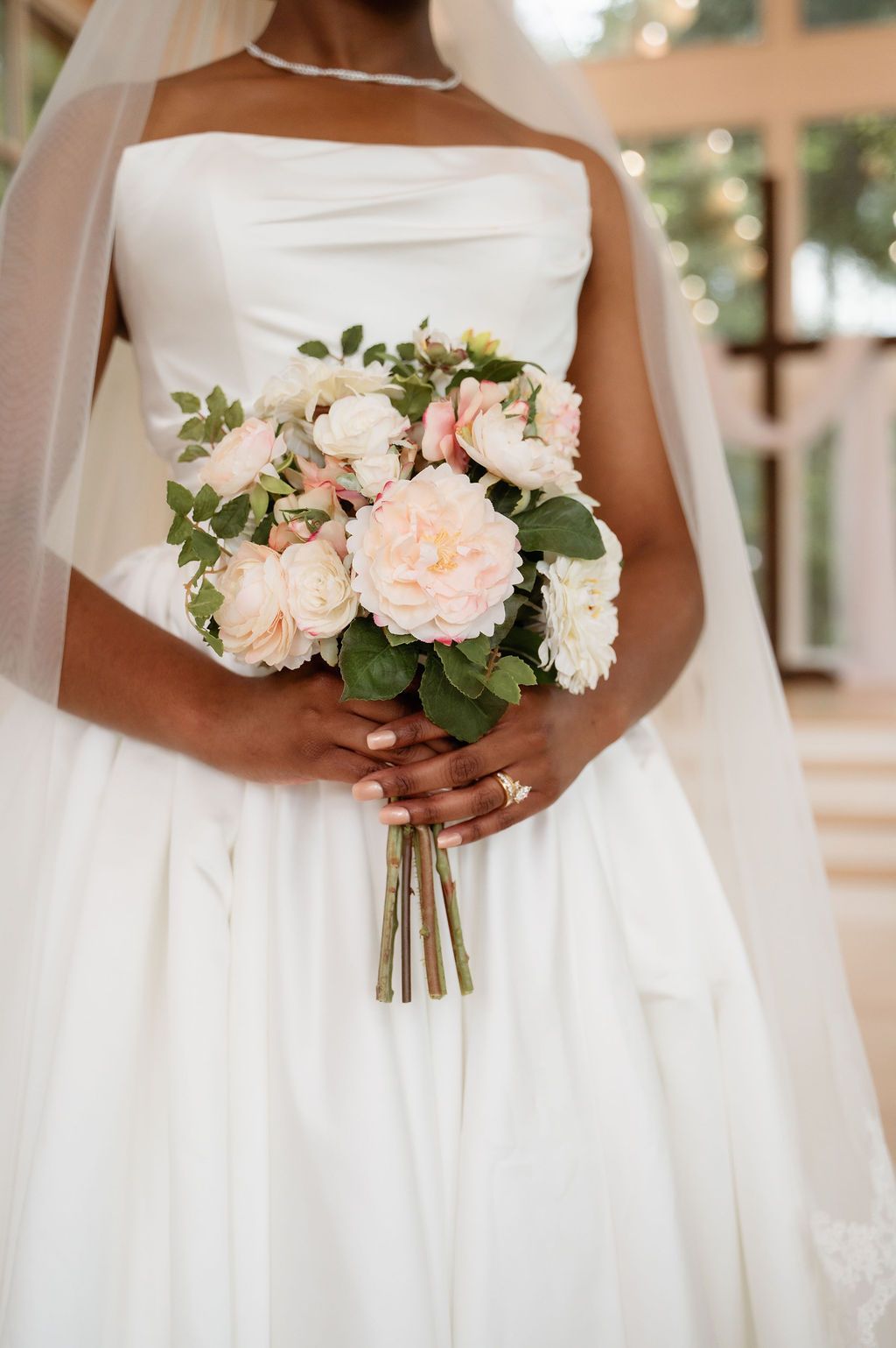 Bride in strapless white gown holding elegant blush and cream peony bouquet with greenery on her wedding day.