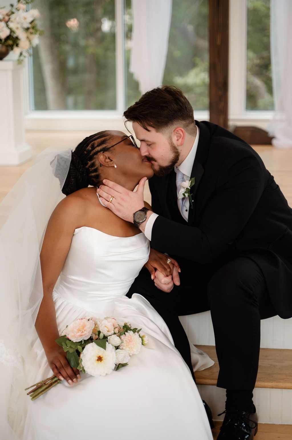 Interracial couple shares a romantic kiss on their wedding day, bride holding blush bouquet.