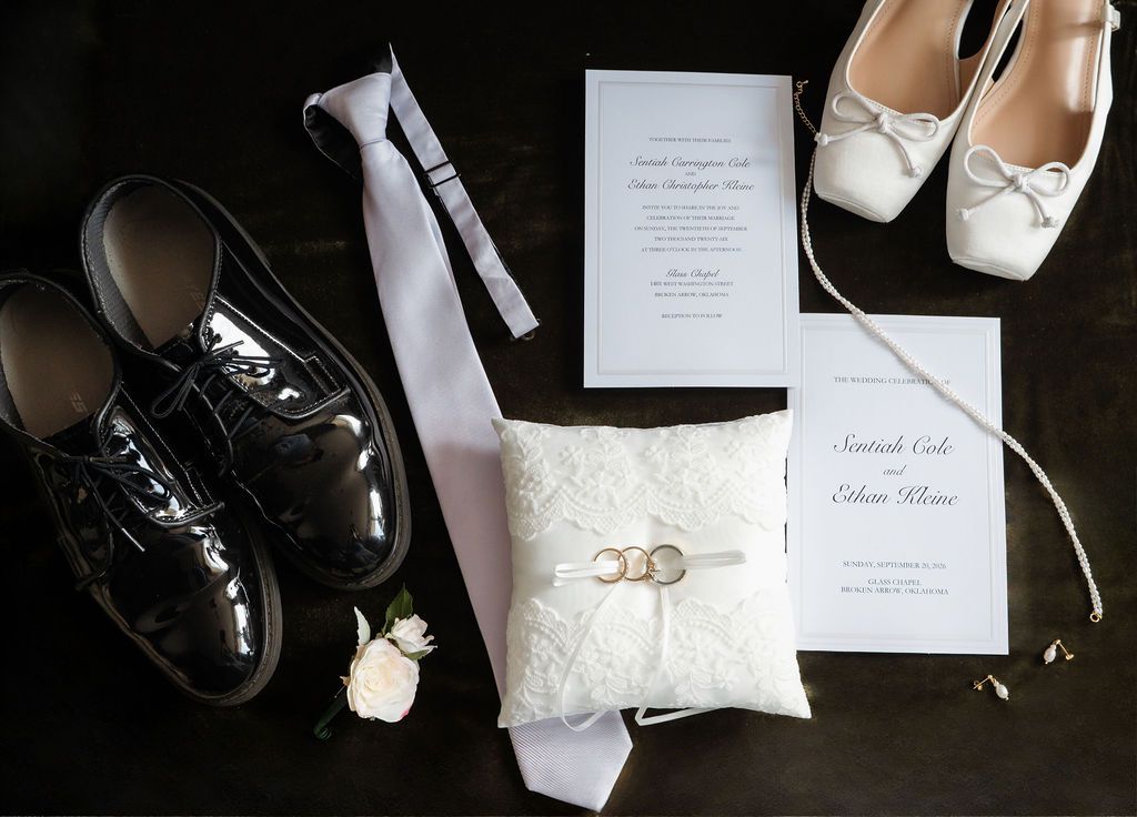Wedding detail flat lay featuring patent leather shoes, white tie, ring pillow with bands, heels, and invitation suite.