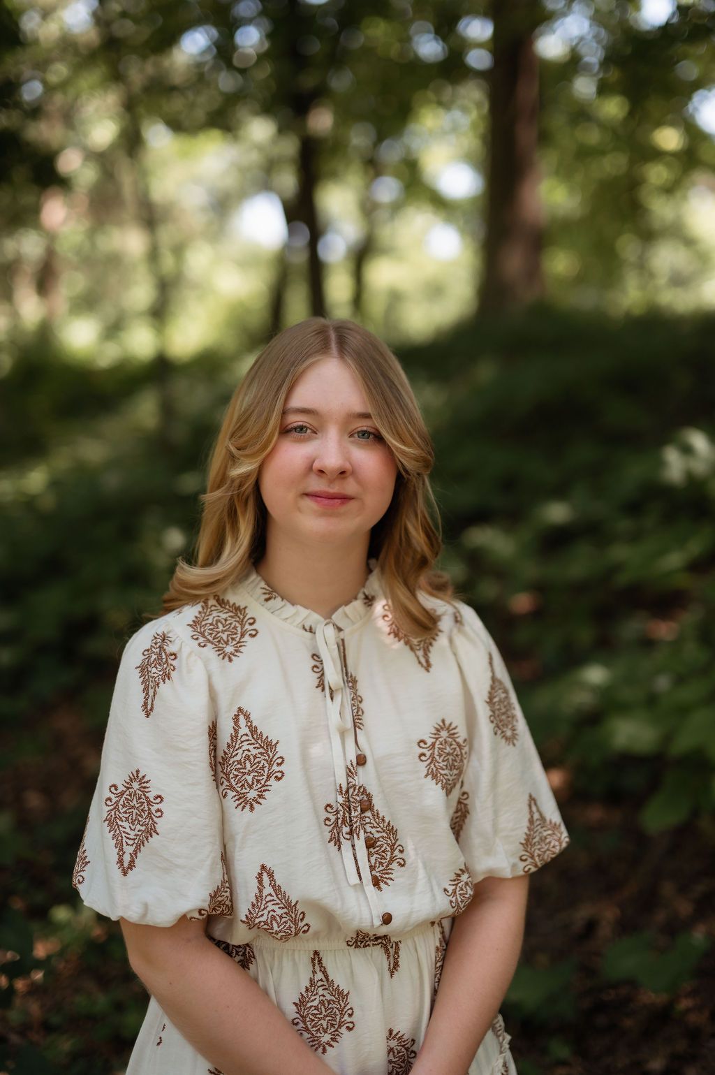 Young woman in cream paisley dress poses in lush green forest for senior portrait photography.