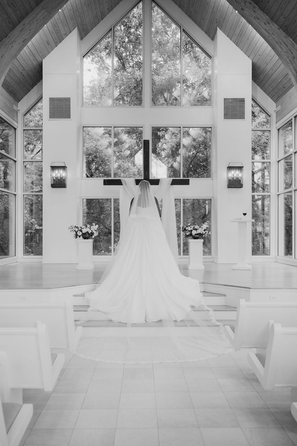 Black and white photo of a bride in a flowing gown standing at the altar of a sunlit chapel with a large cross.