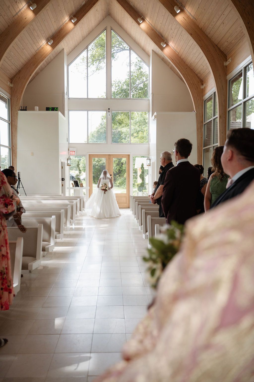 Bride walking down the aisle of a bright chapel with wooden vaulted ceilings and large windows during wedding ceremony.