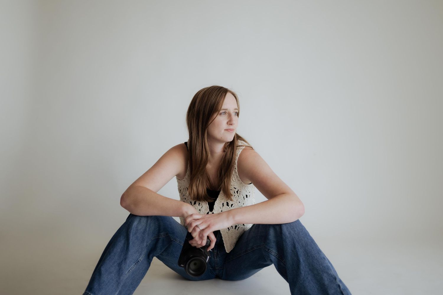 Young woman in jeans and floral top sitting on floor, looking sideways in a minimalist white studio setting.