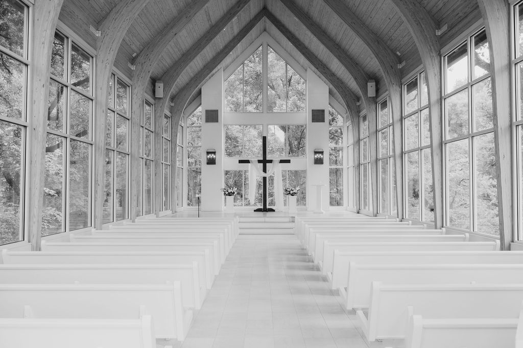 Black and white interior of a modern glass chapel with white pews and a cross at the altar.