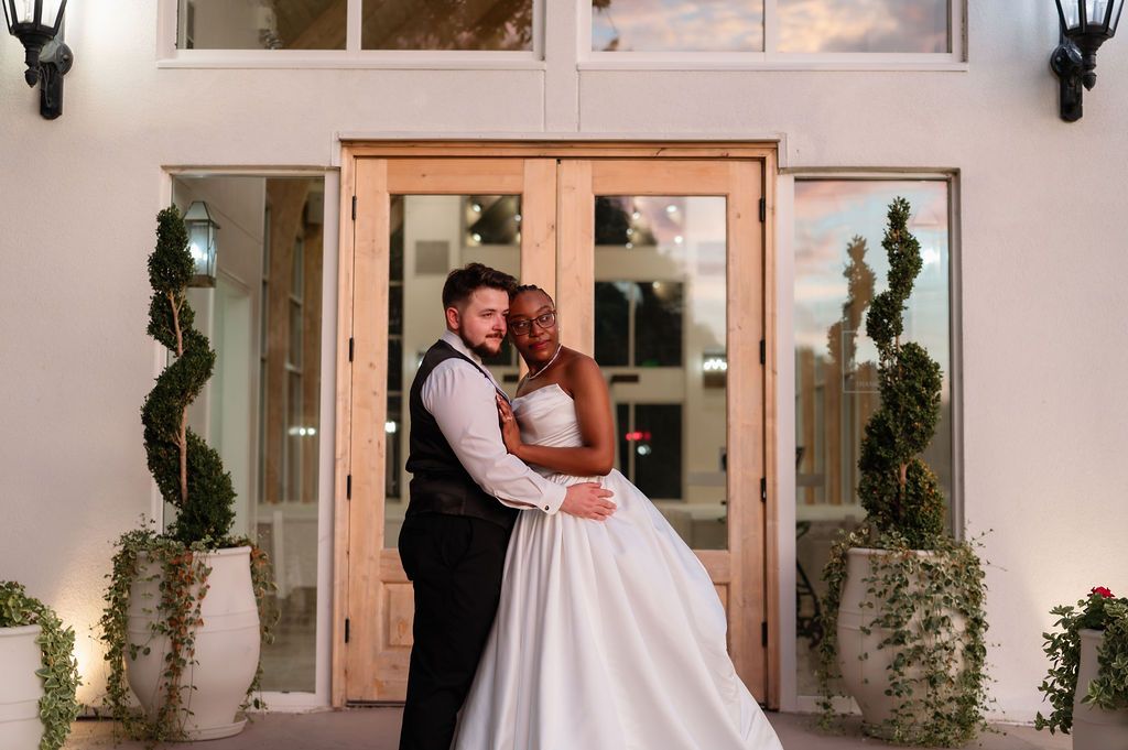 Interracial couple in wedding attire embracing before elegant venue doors with topiary plants at dusk.