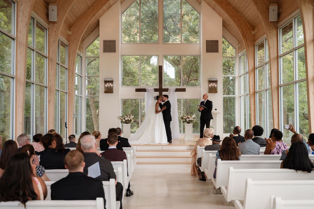 Elegant wedding ceremony in a glass chapel with wooden arches, couple at altar, guests seated in white pews.