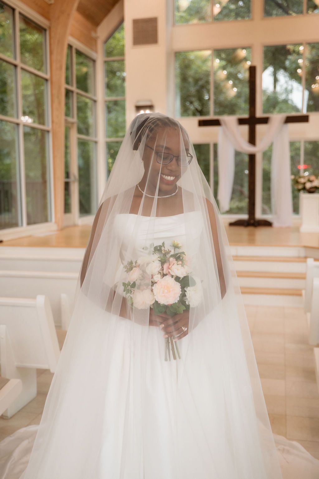 Smiling bride in white gown and veil holds bouquet in sunlit chapel with cross backdrop.