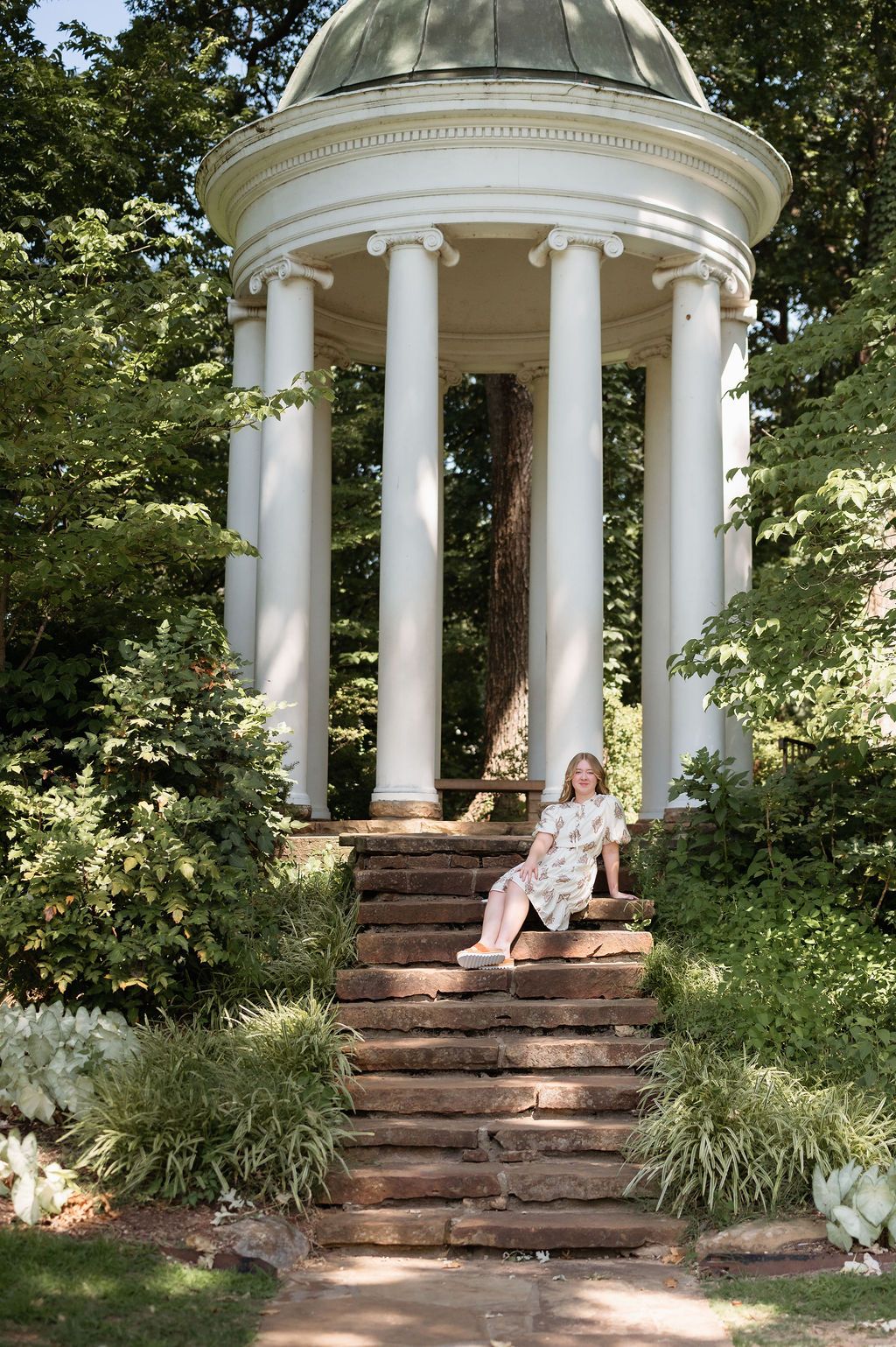Young woman in floral dress sits on stone steps of elegant white columned garden rotunda.