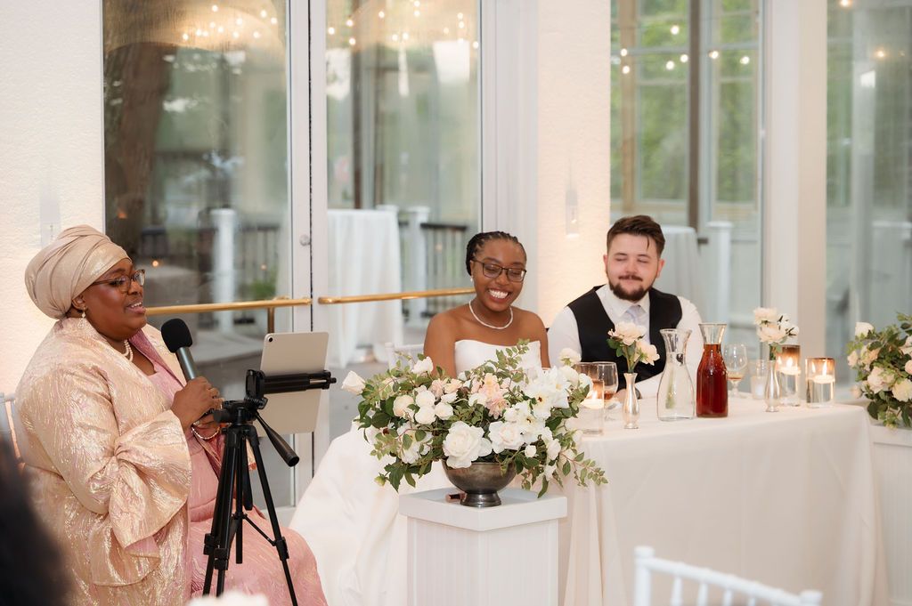 A joyful wedding reception with a guest speaking at a microphone while the bride and groom smile at the sweetheart table.