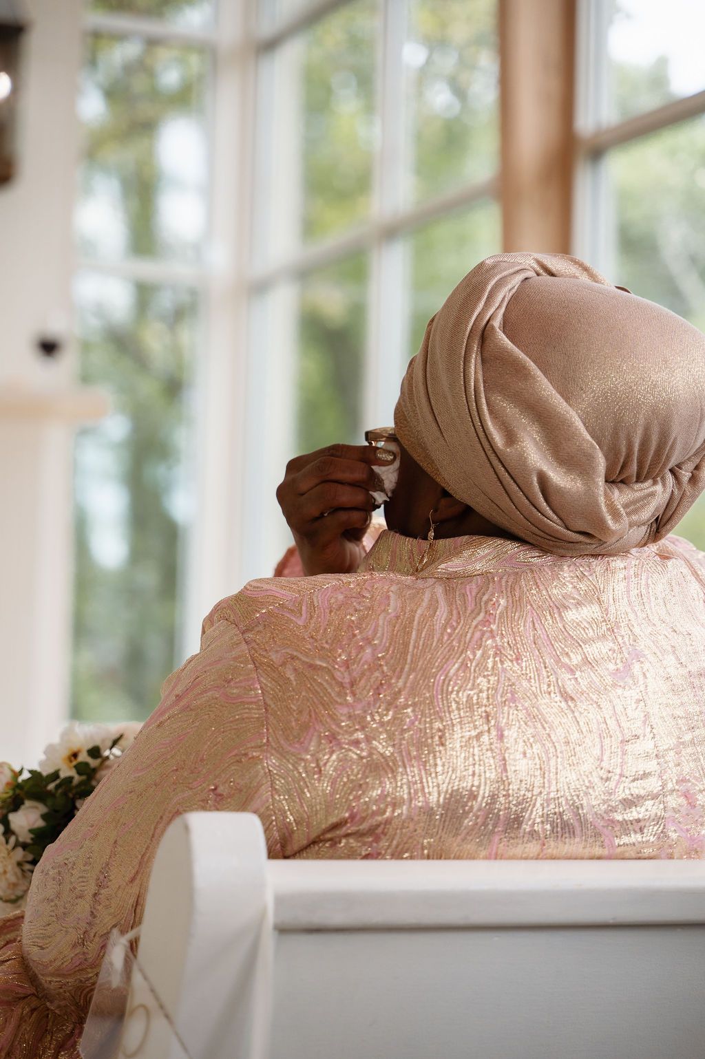 Elegant woman in rose gold shimmer dress and matching turban headwrap seated by bright chapel windows.