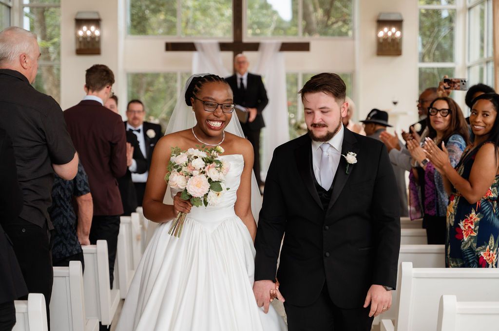 Joyful interracial newlywed couple walks down church aisle, bride in white gown holding pink bouquet, groom in black suit.