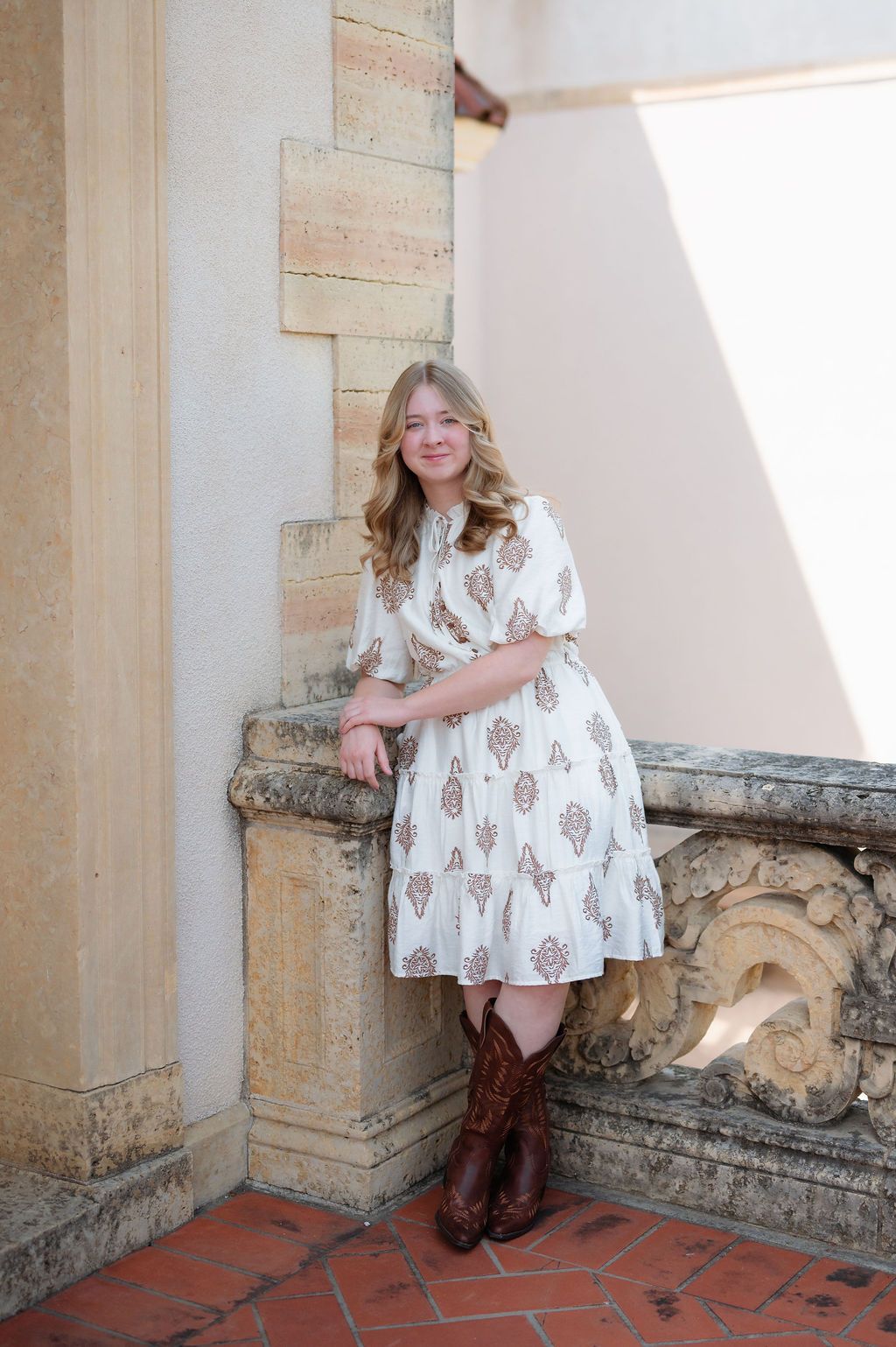 Smiling young woman in white patterned dress and brown cowboy boots leans against stone balustrade.