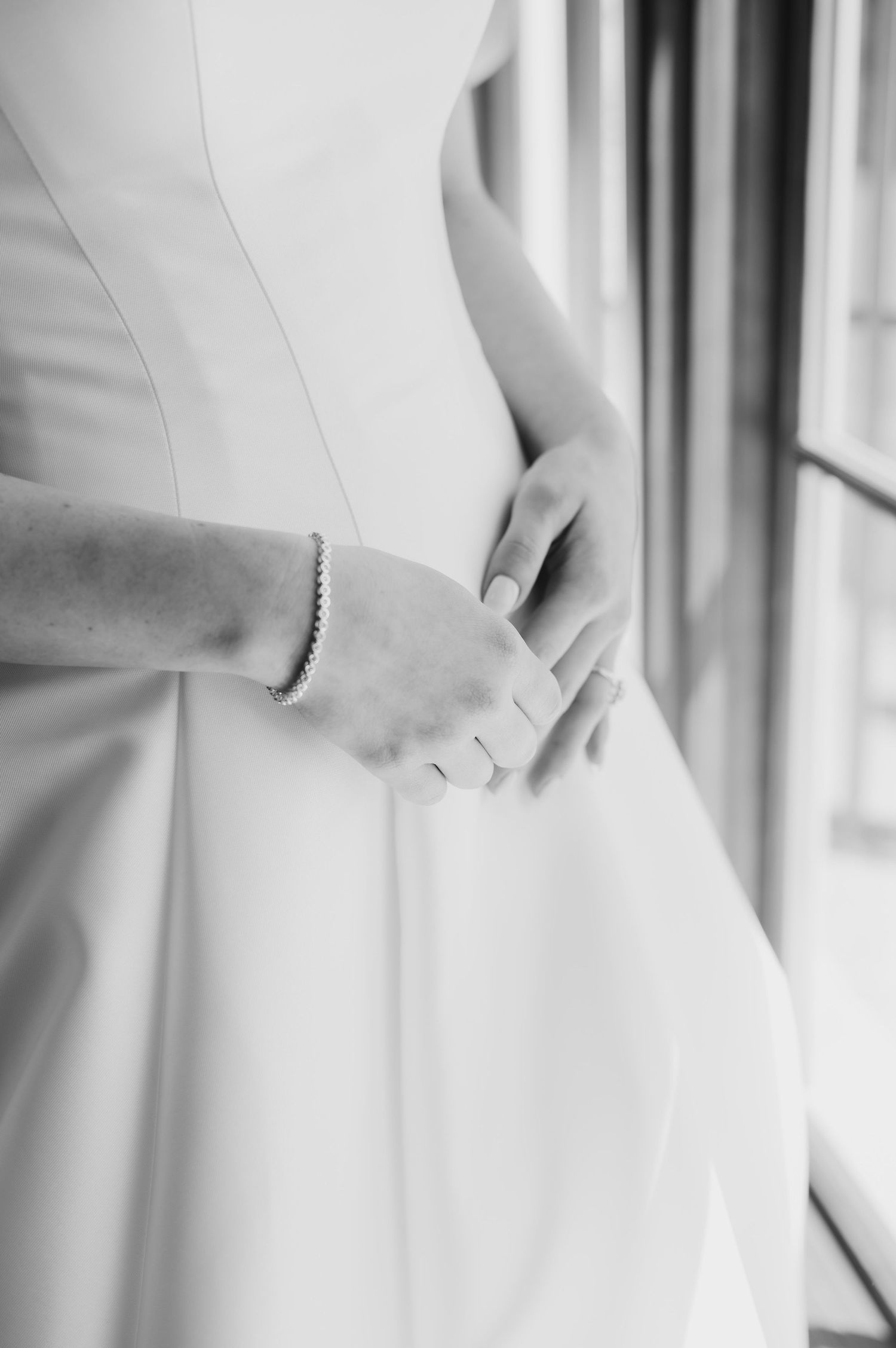 Black and white photo of a bride in a white gown clasping her hands at her waist, wearing a delicate bracelet.