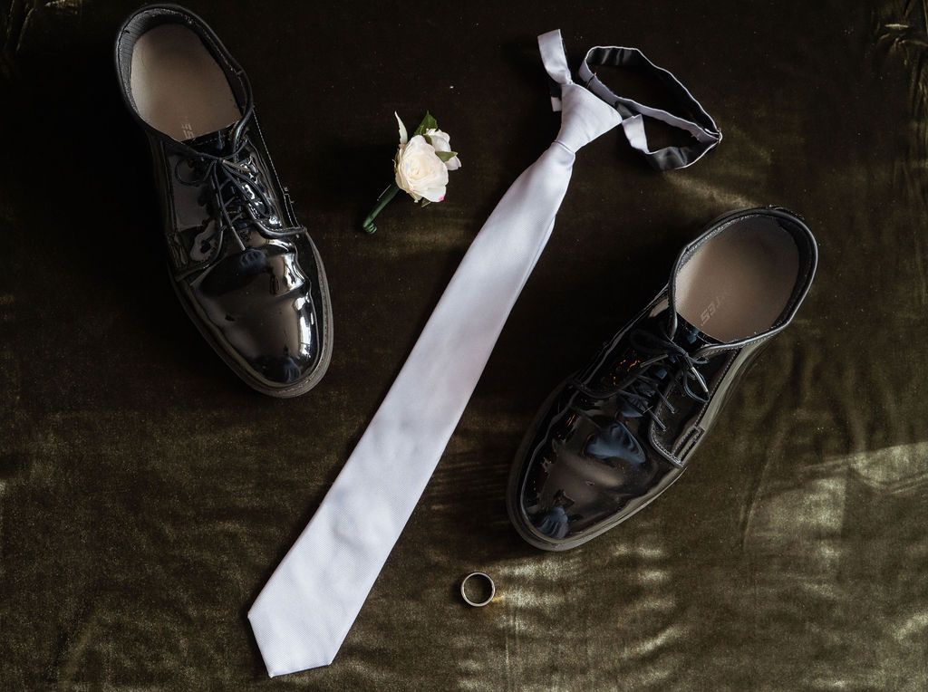 Black patent leather dress shoes, white tie, boutonniere, and gold ring arranged on dark surface for wedding.