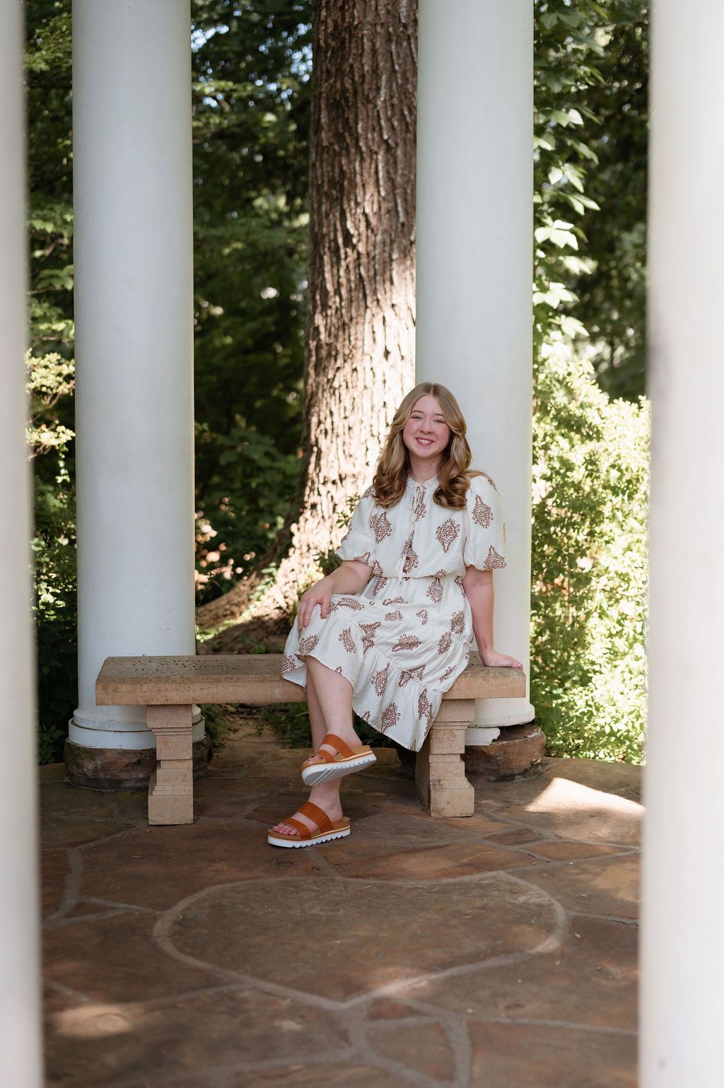 Smiling young woman in cream floral dress sits on bench between white columns for senior photos.