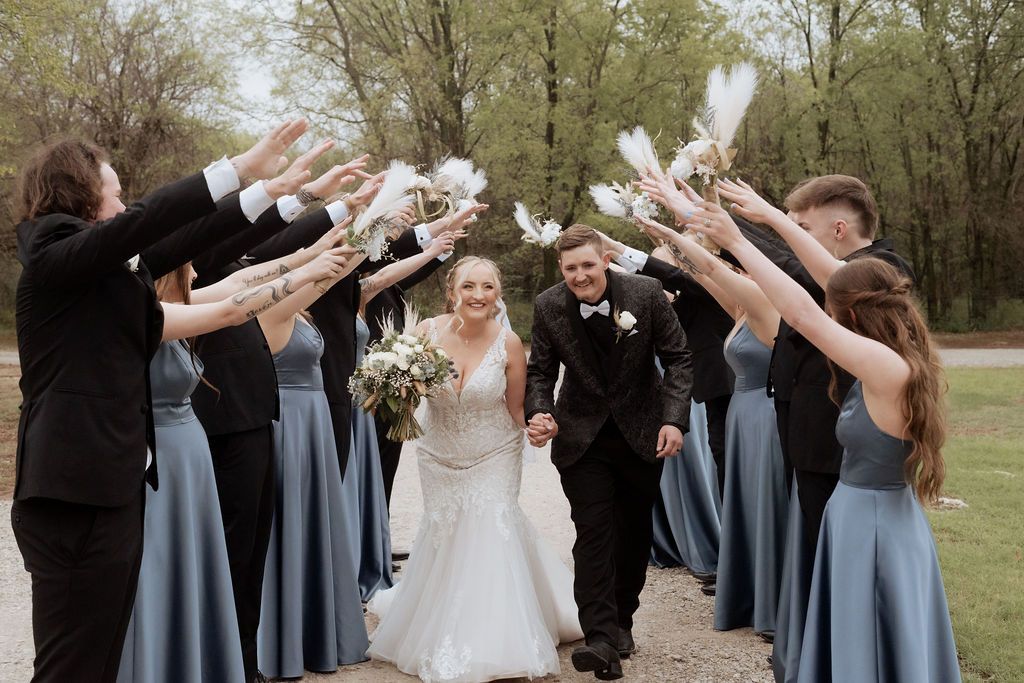 Bride and groom walk through arch of raised hands held by wedding party in dusty blue dresses and black suits outdoors.