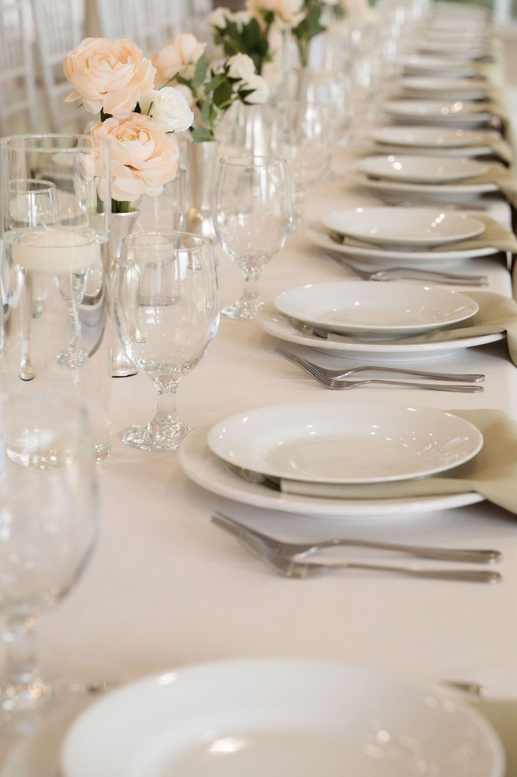 Elegant wedding reception table setting with white plates, crystal glasses, silver cutlery, and blush pink rose centerpieces.