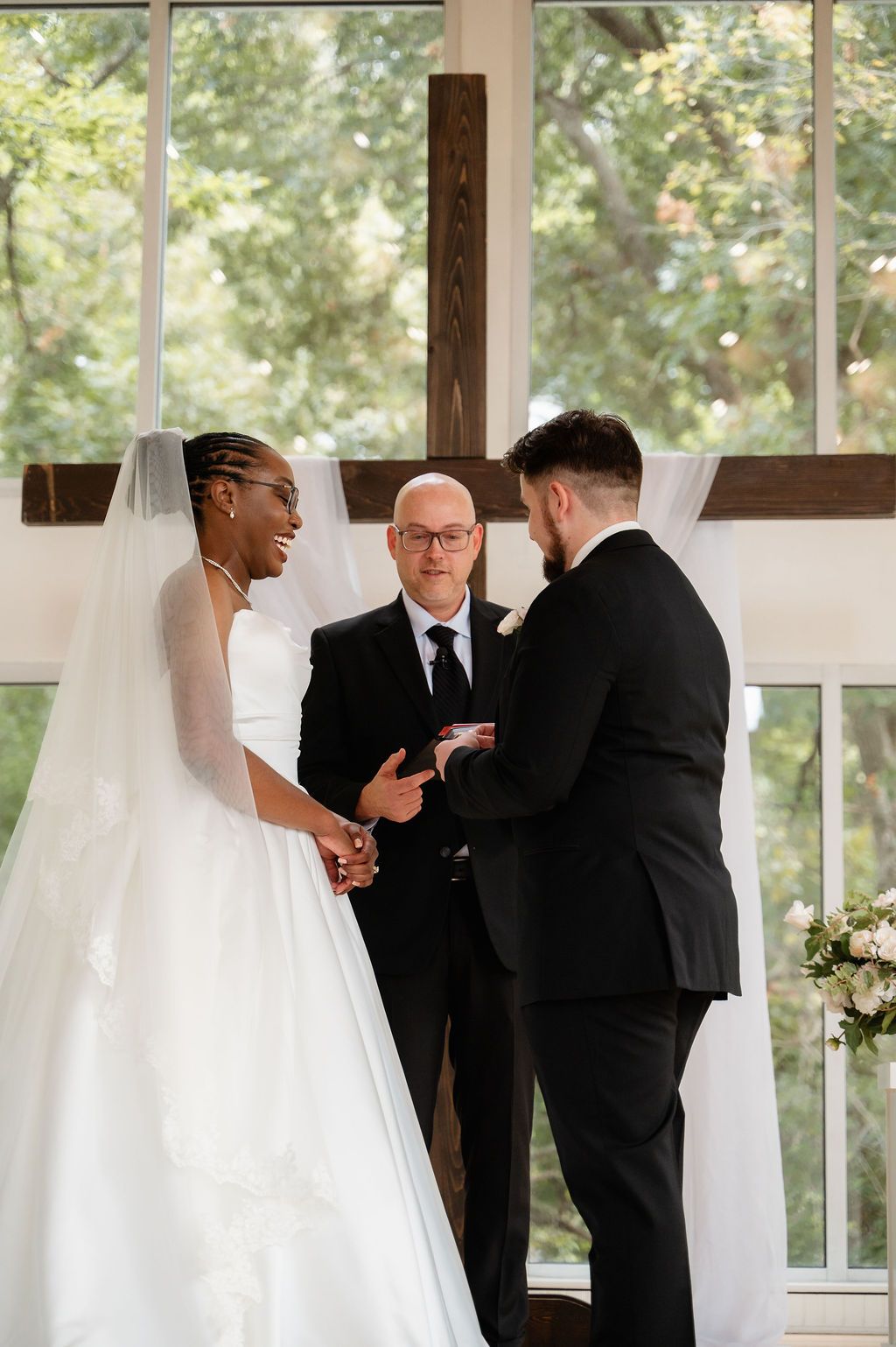 Interracial couple exchanging vows at chapel altar with officiant and wooden cross backdrop.