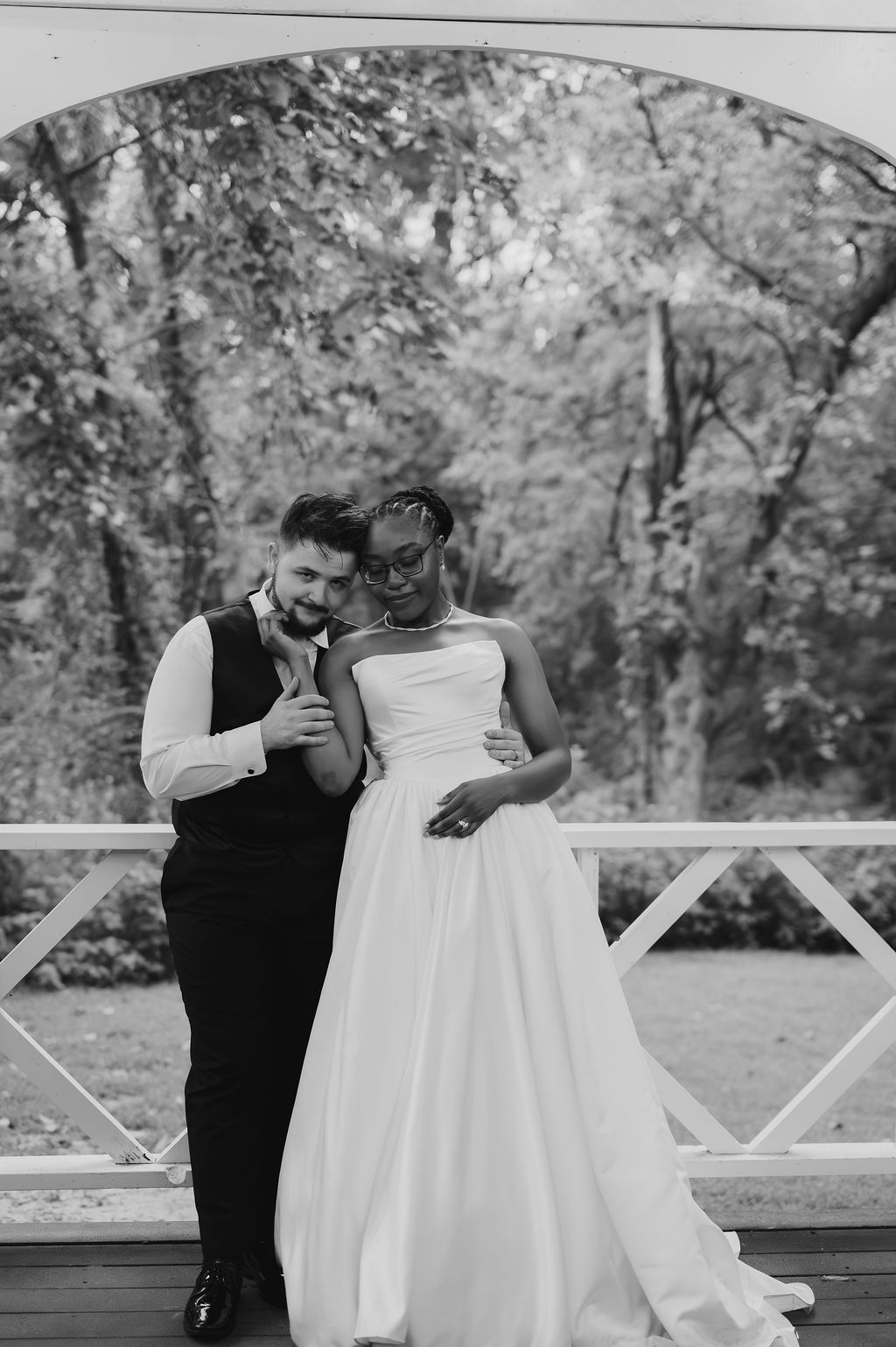 Black and white portrait of interracial newlyweds posing together on outdoor bridge, bride in strapless gown, groom in vest.